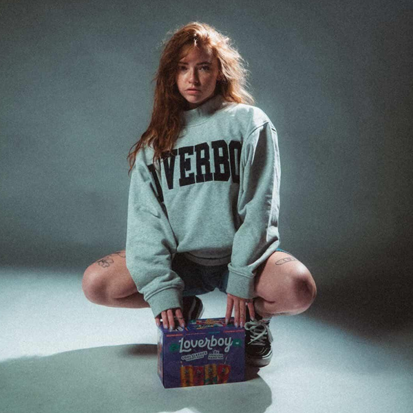 A woman crouches on a studio floor wearing a light gray sweatshirt with 'LOVERBOY' printed in black across the chest, holding a purple 'Lovenboy' box. The background is a dark gray studio backdrop.