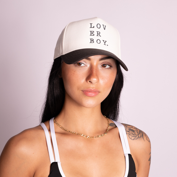 A woman wearing a natural and black trucker cap with the text 'LOVER BOY.' printed vertically in black on the front, set against a solid pale pink background.
