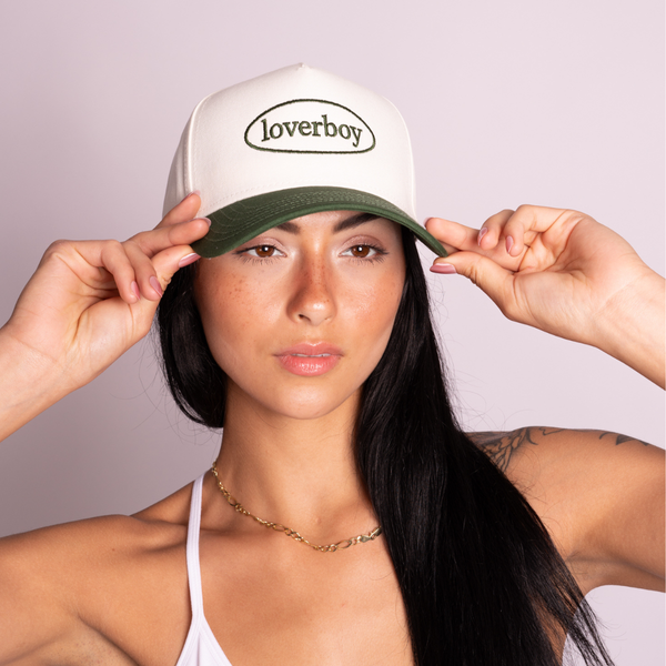A woman wearing a off white and forest green five-panel style cap with the brand name 'loverboy' embroidered on the front, holding the brim with both hands against a light lavender background.